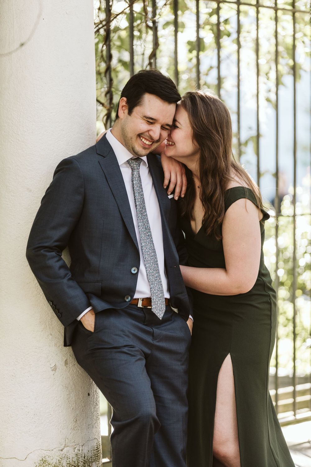 A couple shares a romantic moment, woman in green dress whispering to smiling man in navy suit leaning against white column.