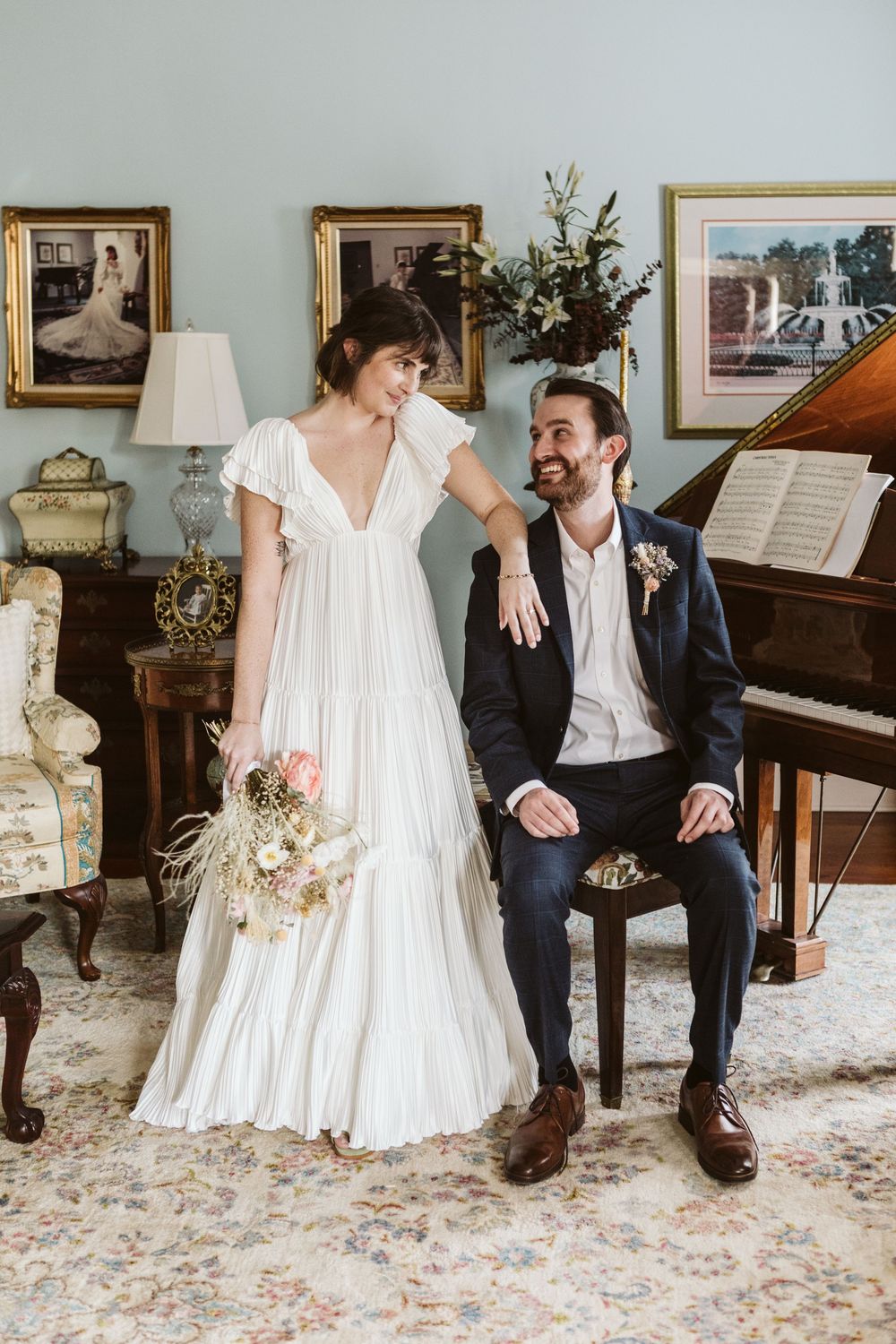 Bride in white dress holding bouquet stands beside groom in navy suit near a grand piano in an elegant room.