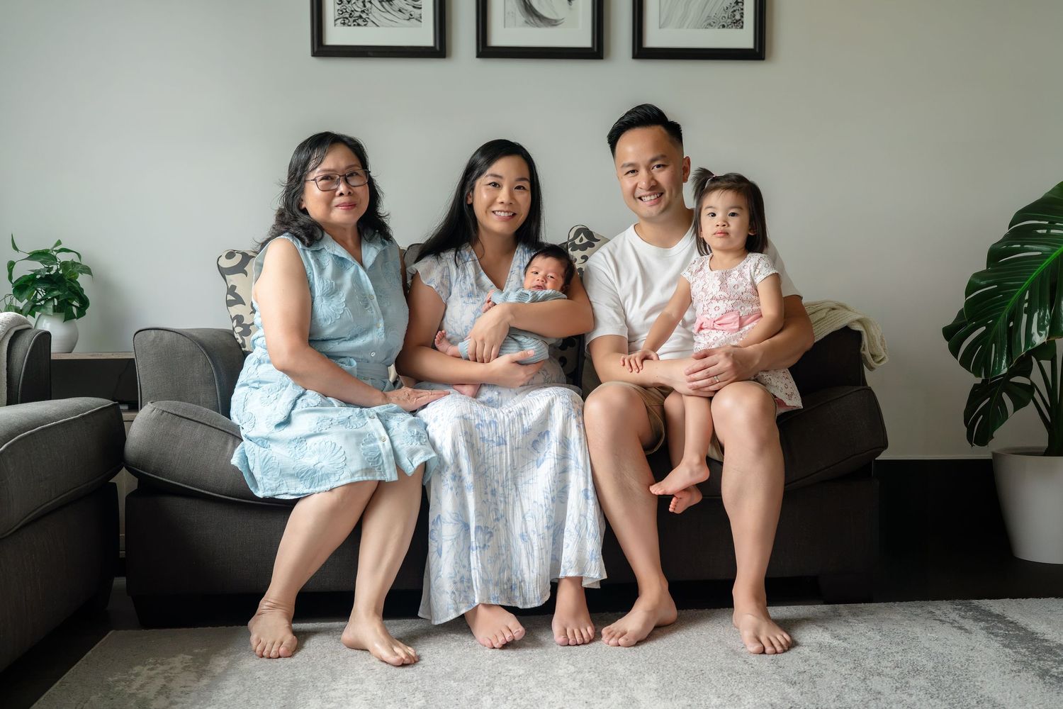 Multi-generational Asian family portrait with grandmother, parents, toddler, and newborn baby sitting together on a couch.