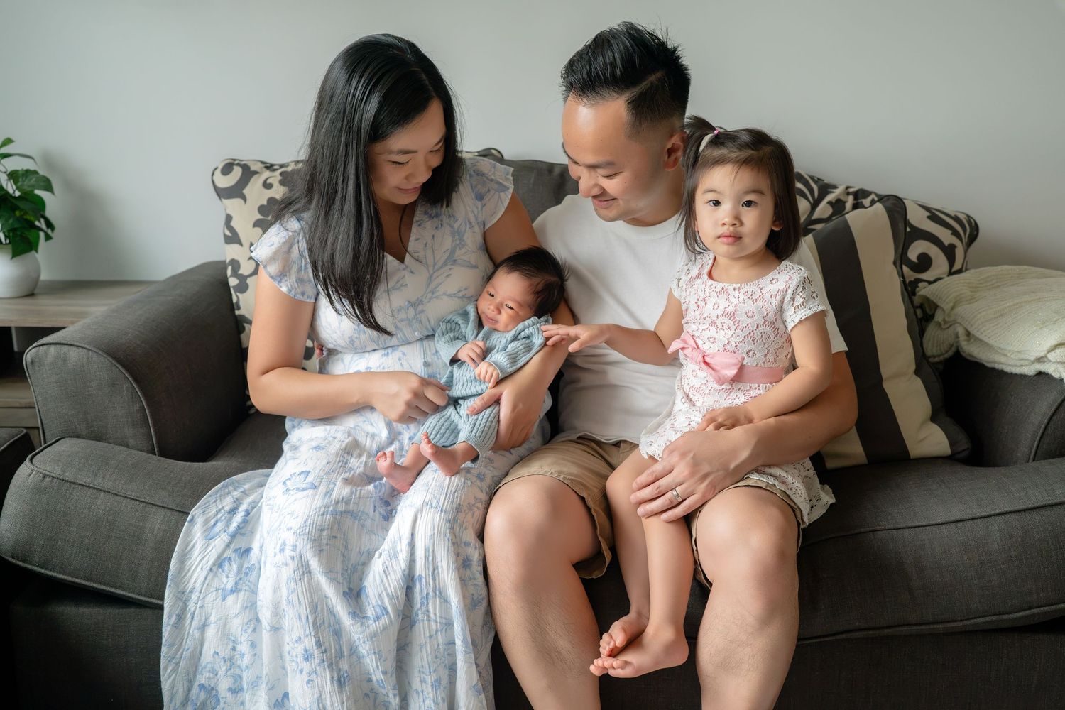 A happy Asian family of four sitting on a gray sofa, parents holding a newborn baby and toddler daughter at home.