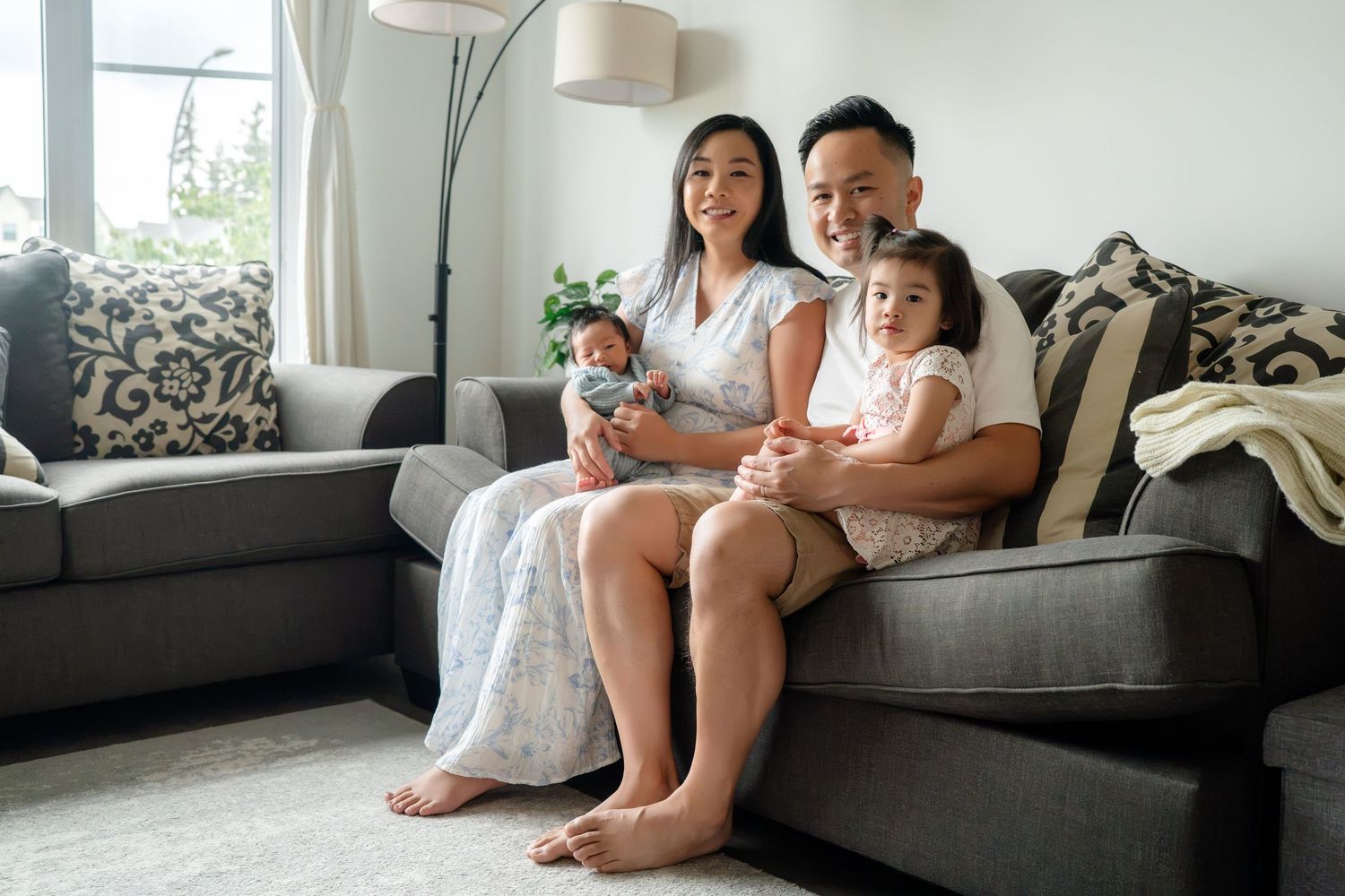 Happy Asian family of four sitting on a gray sofa, parents holding two young children in a bright modern living room.