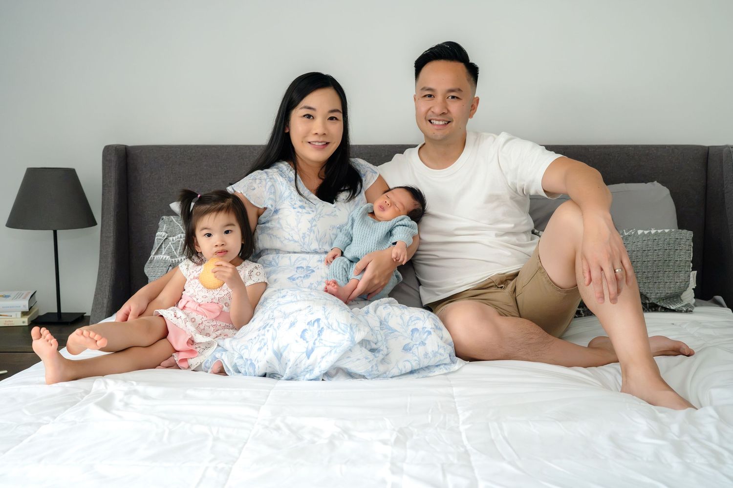 Happy Asian family of four sitting on a white bed, parents smiling with toddler girl and newborn baby.
