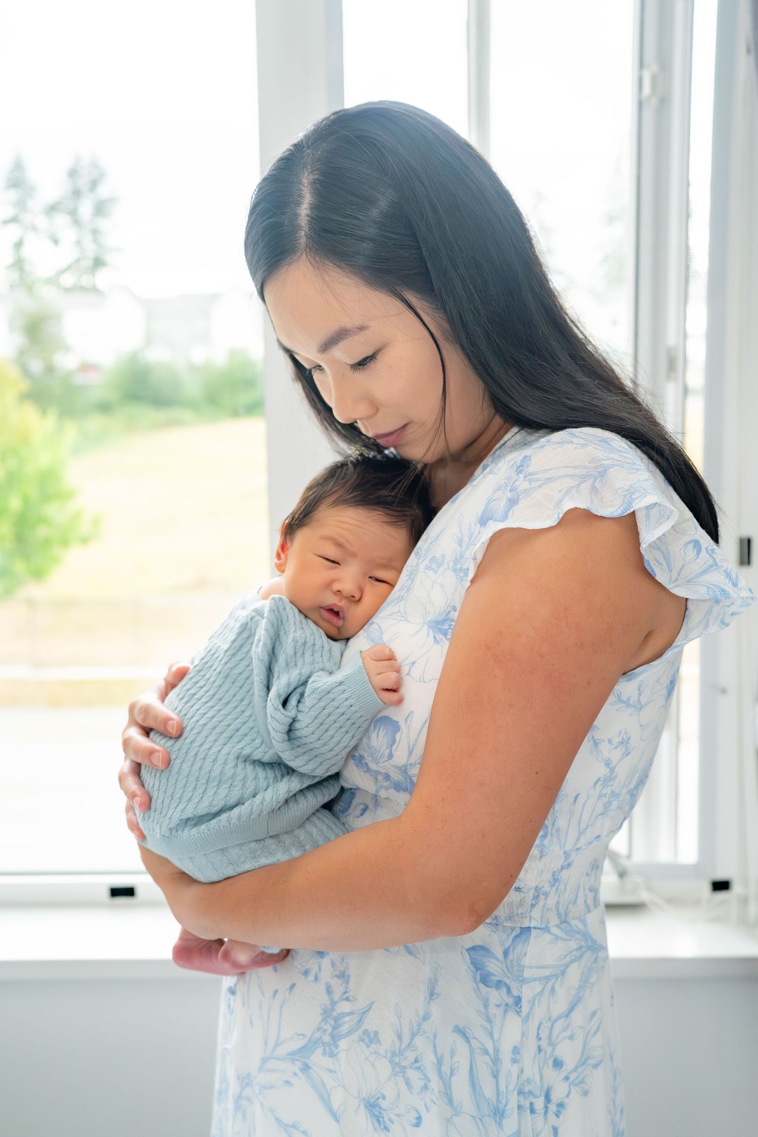 Young Asian mother in floral dress tenderly holds sleeping newborn baby near bright window indoors.