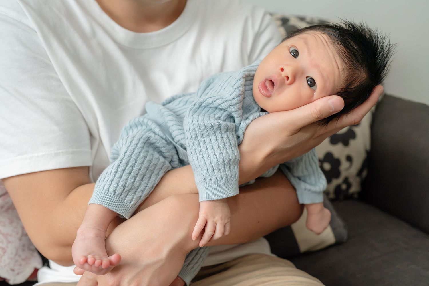 Parent gently cradling a newborn baby in blue knit outfit on a couch.