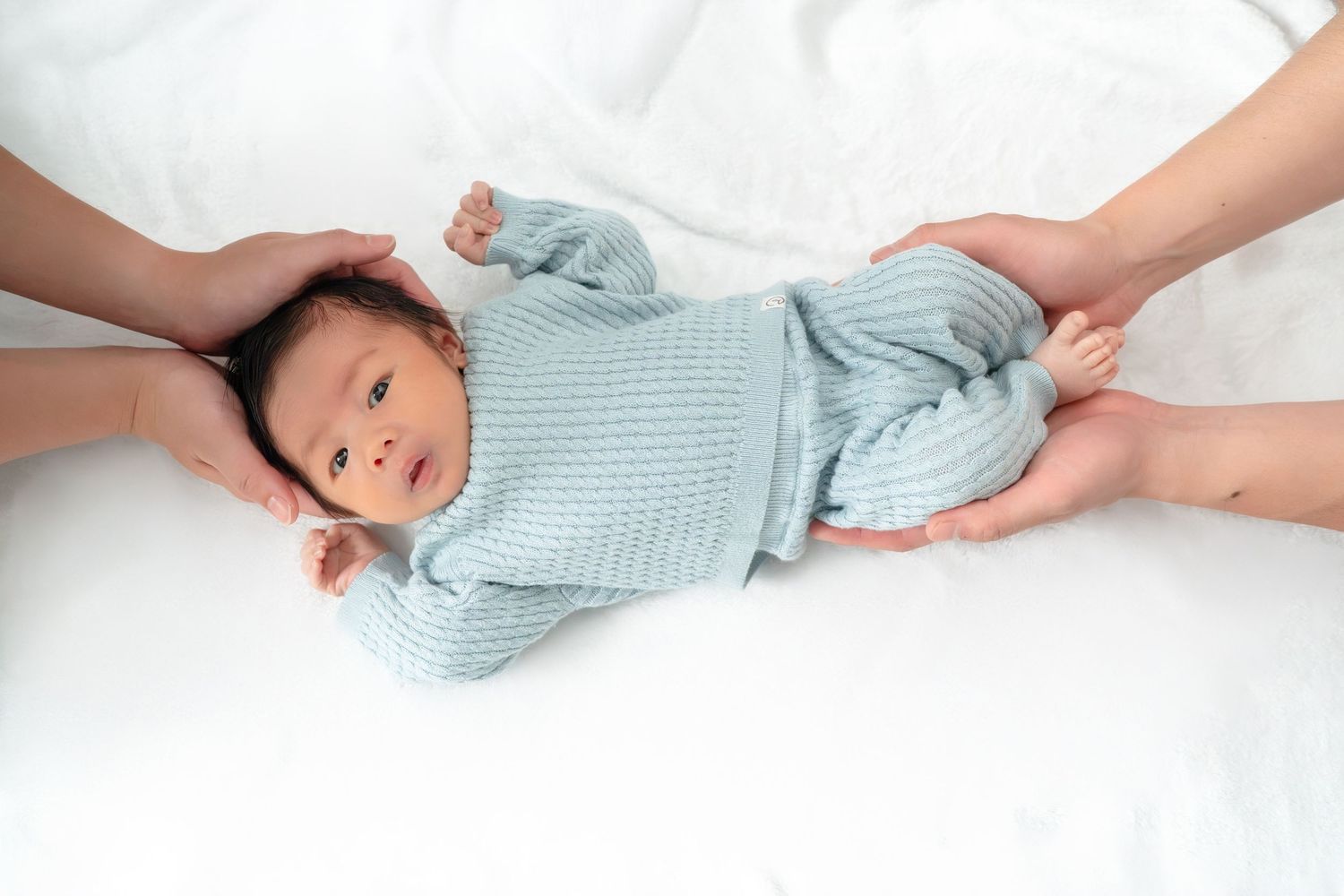 Newborn baby in light blue knit outfit lying on white blanket while two adults gently hold the infant.