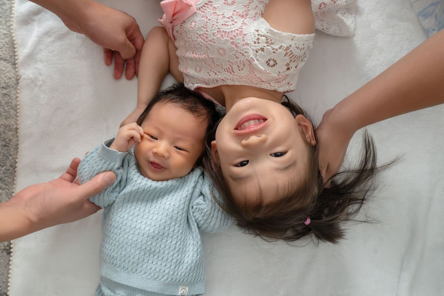 Two young Asian siblings, a newborn baby and toddler girl, lying together on a white blanket held by adult hands.