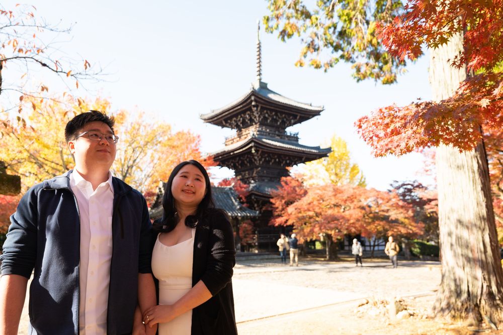 Smiling couple poses together in front of a traditional Japanese pagoda surrounded by vibrant red and orange autumn maple leaves.