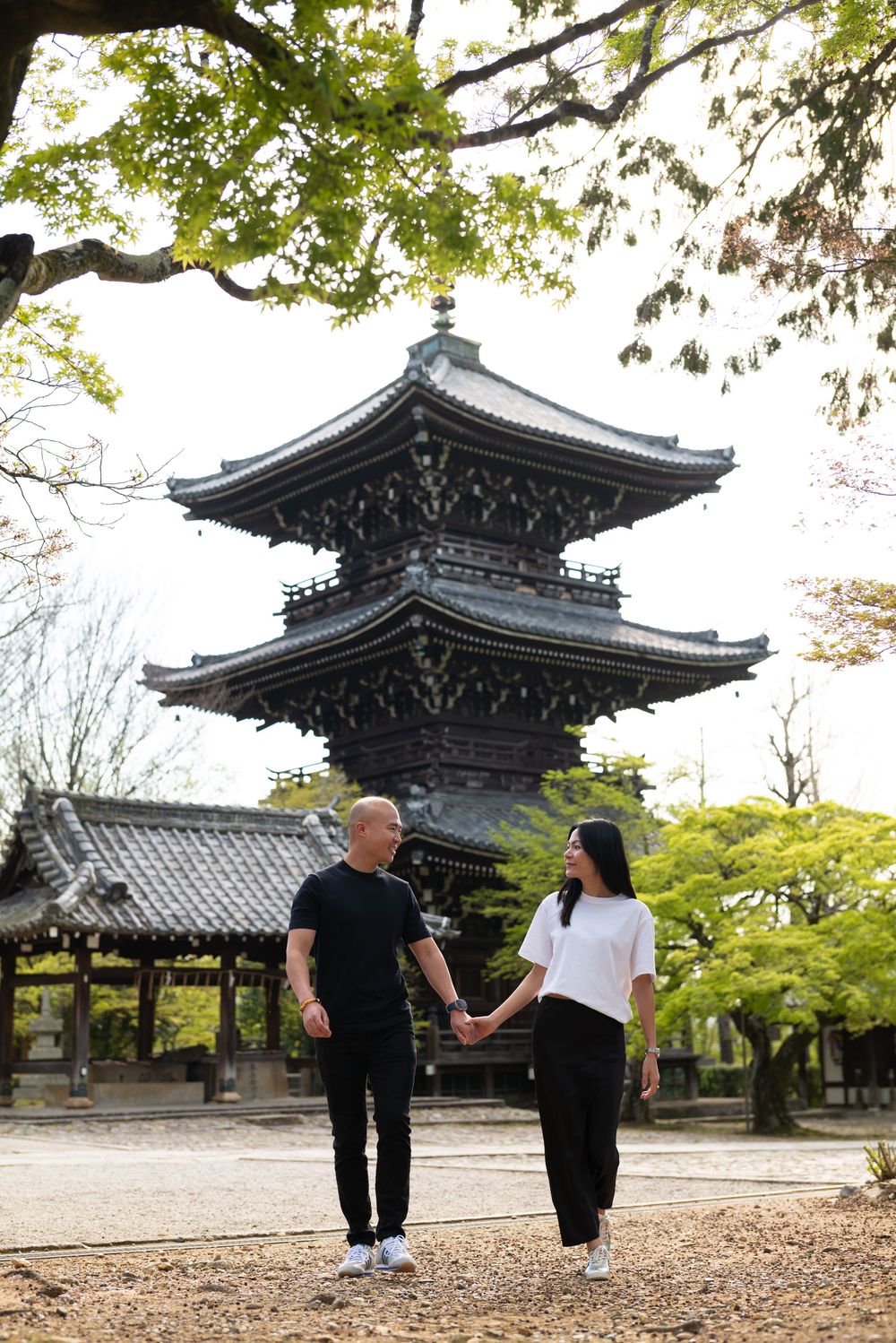 Couple in black and white outfits holding hands and walking past a traditional Japanese wooden pagoda surrounded by green trees.