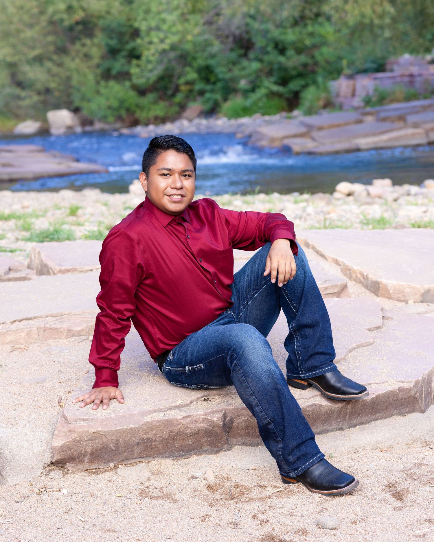 Young man in red shirt and jeans sits relaxed on sandstone rocks near a flowing river in an outdoor portrait.