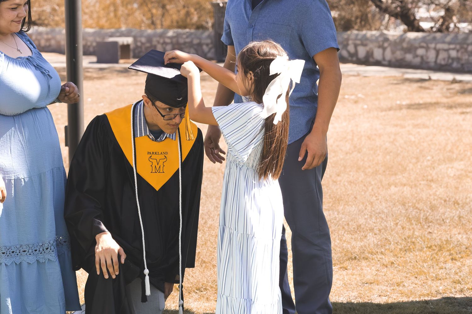 A girl adjusts a graduate’s cap as family gathers around him outdoors in a candid graduation moment.