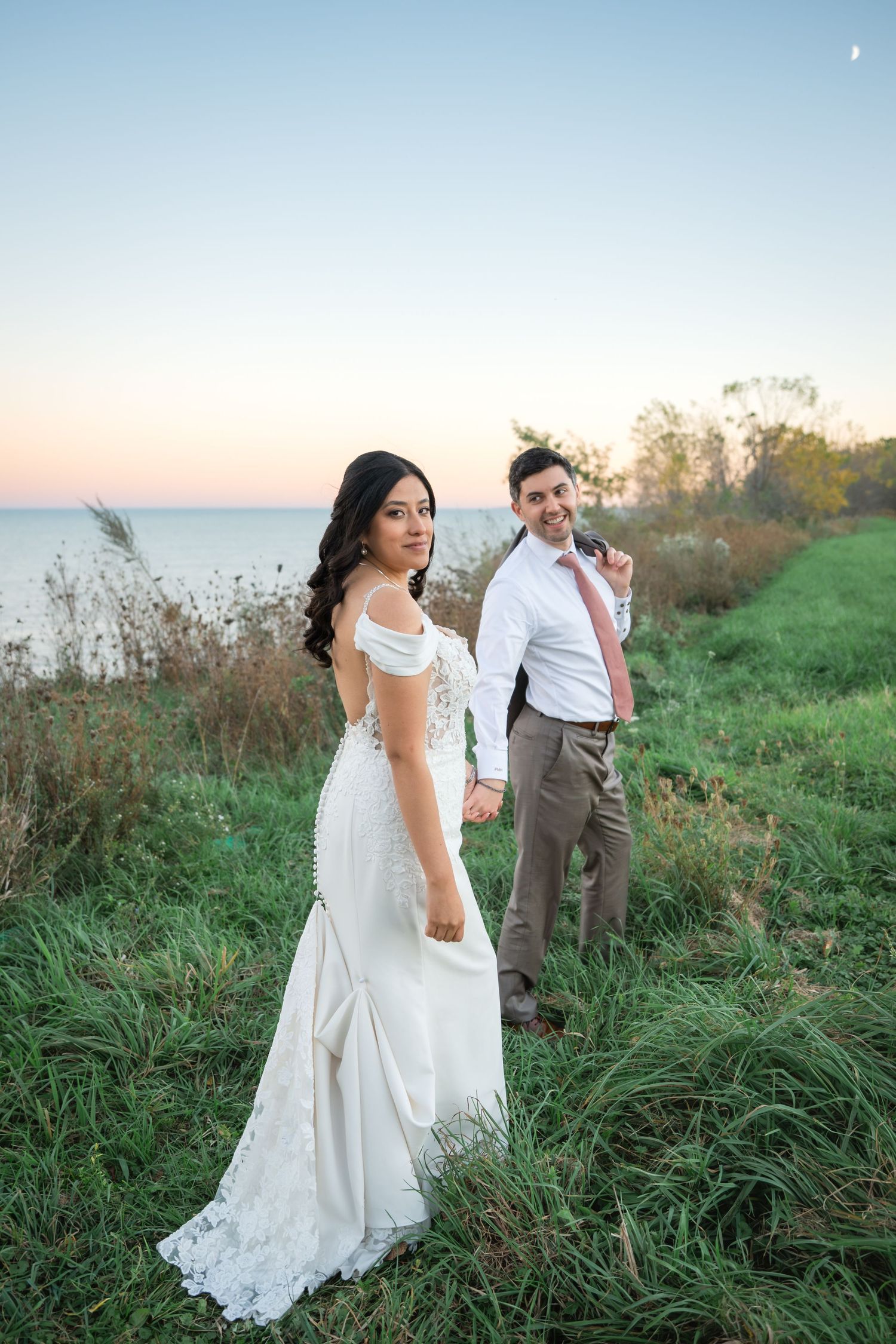 Bride in a white lace wedding gown holds hands with a smiling groom in a pink tie while walking through a grassy lakeside field at sunset.