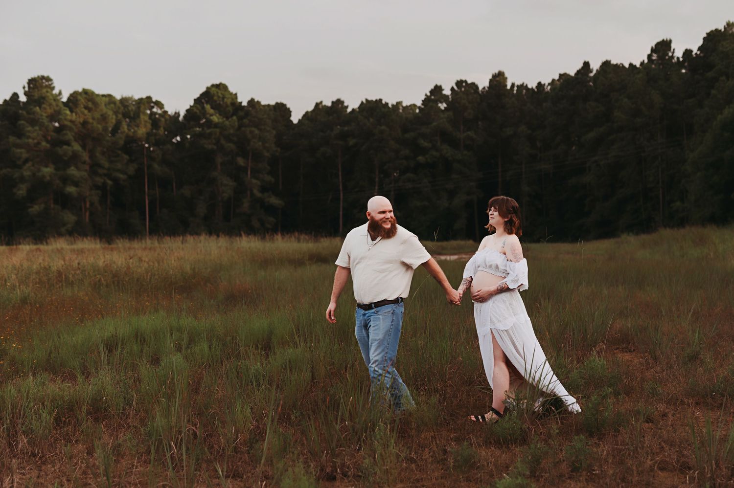 Smiling couple holding hands and walking through a grassy meadow with tall pine trees behind them during a maternity photo session.