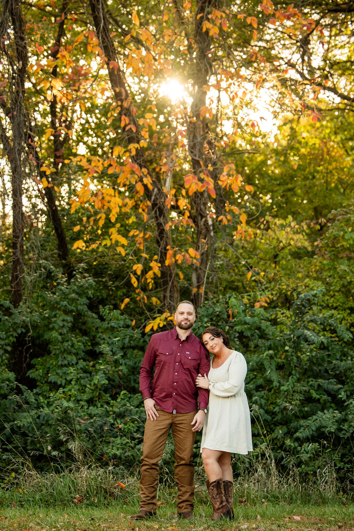 Couple poses together in autumn forest with golden sunlight filtering through colorful fall foliage.