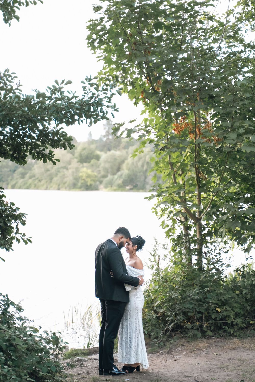 Bride in a white gown and groom in a dark suit embrace forehead to forehead on a wooded lakeside path surrounded by green trees.