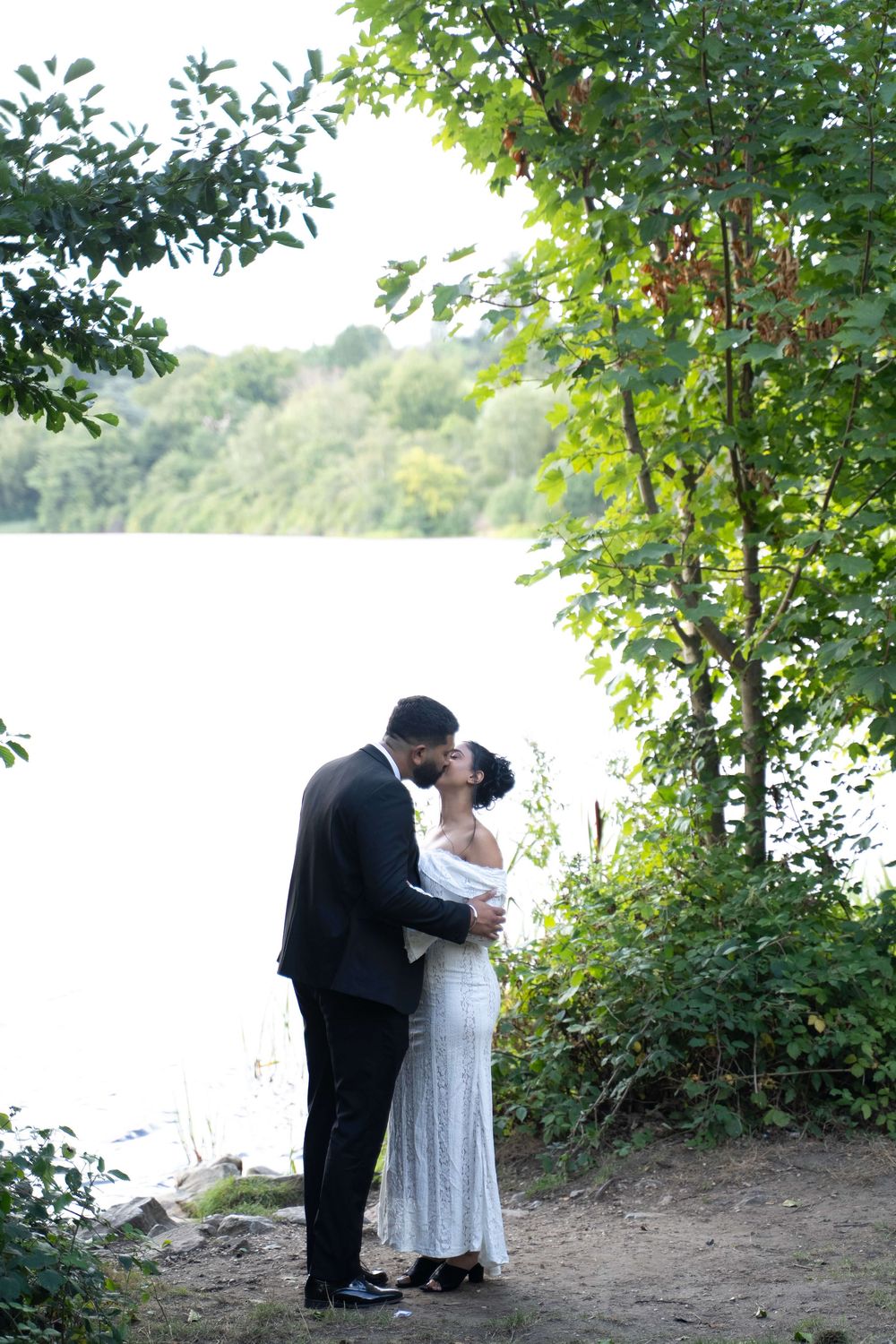 Bride in a white lace gown and groom in a dark suit share a kiss beside a calm lake, framed by green trees.