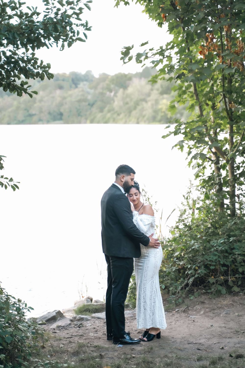 Couple in formal attire embracing on a rocky lakeside shore surrounded by green trees, in an intimate engagement or wedding portrait.