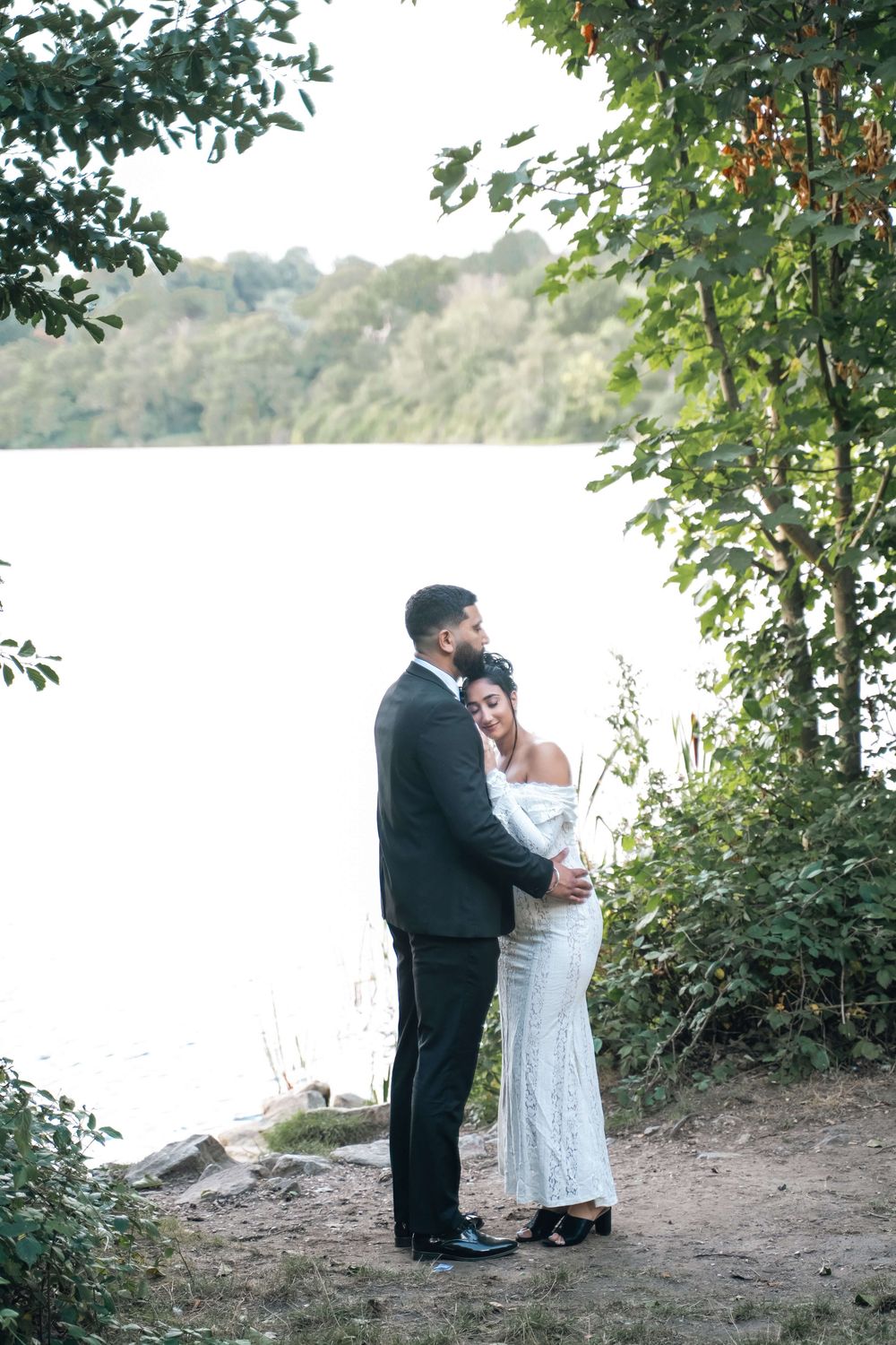 Couple in formal wedding attire embrace on a rocky lakeshore surrounded by green trees during a romantic outdoor photo session.