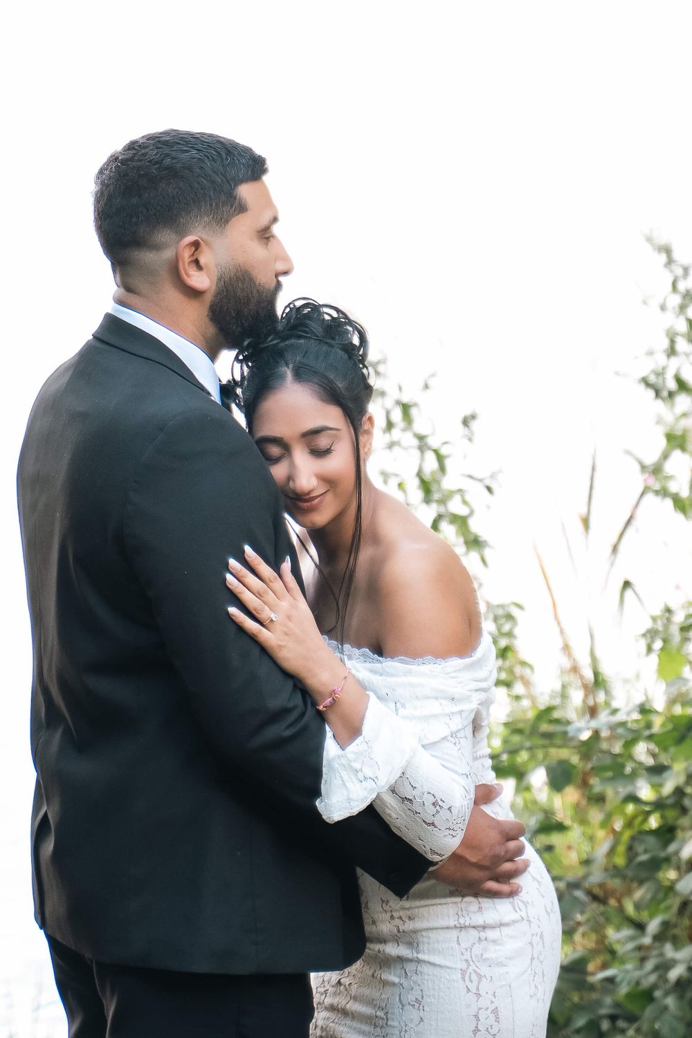 A bearded groom in a dark suit embraces a smiling bride in an off-shoulder white lace gown during an outdoor wedding portrait session.