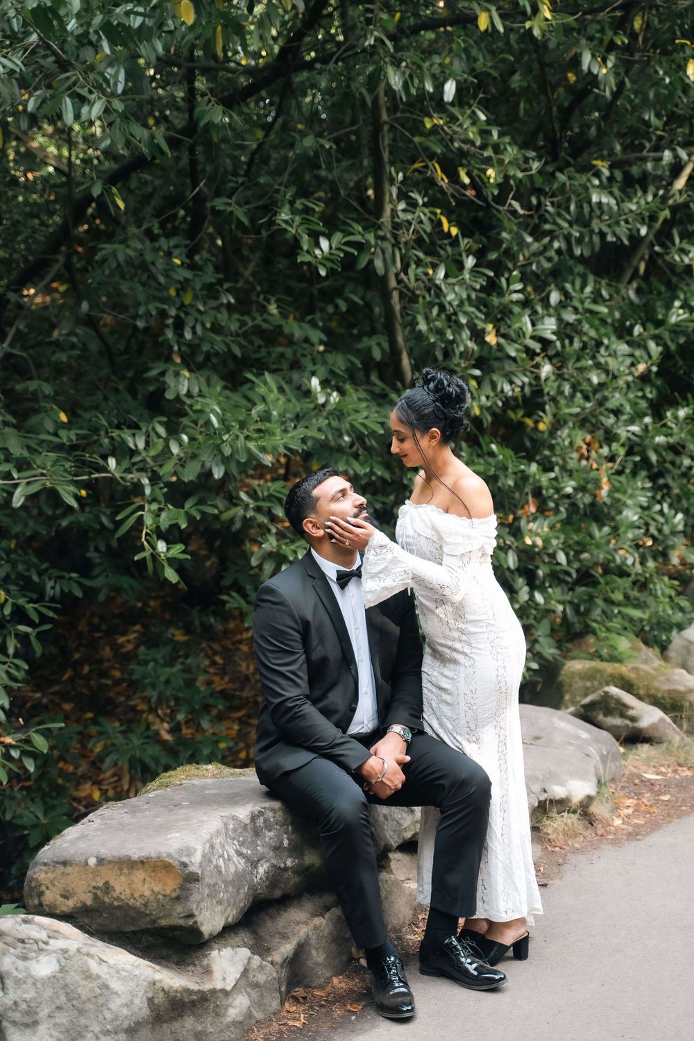 Bride in an off-shoulder white lace gown touches the chin of a seated groom in a black tuxedo on mossy rocks beside a leafy green forest.