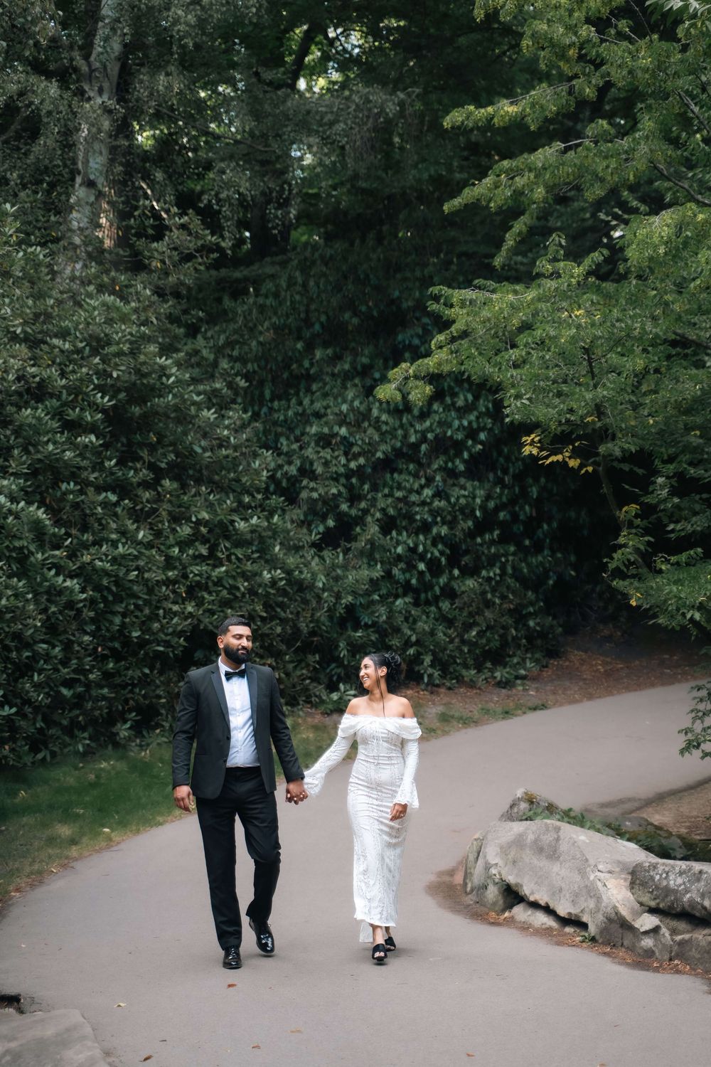 A couple holds hands while walking along a curved park path, the groom in a black tuxedo and the bride in a white off-shoulder gown.