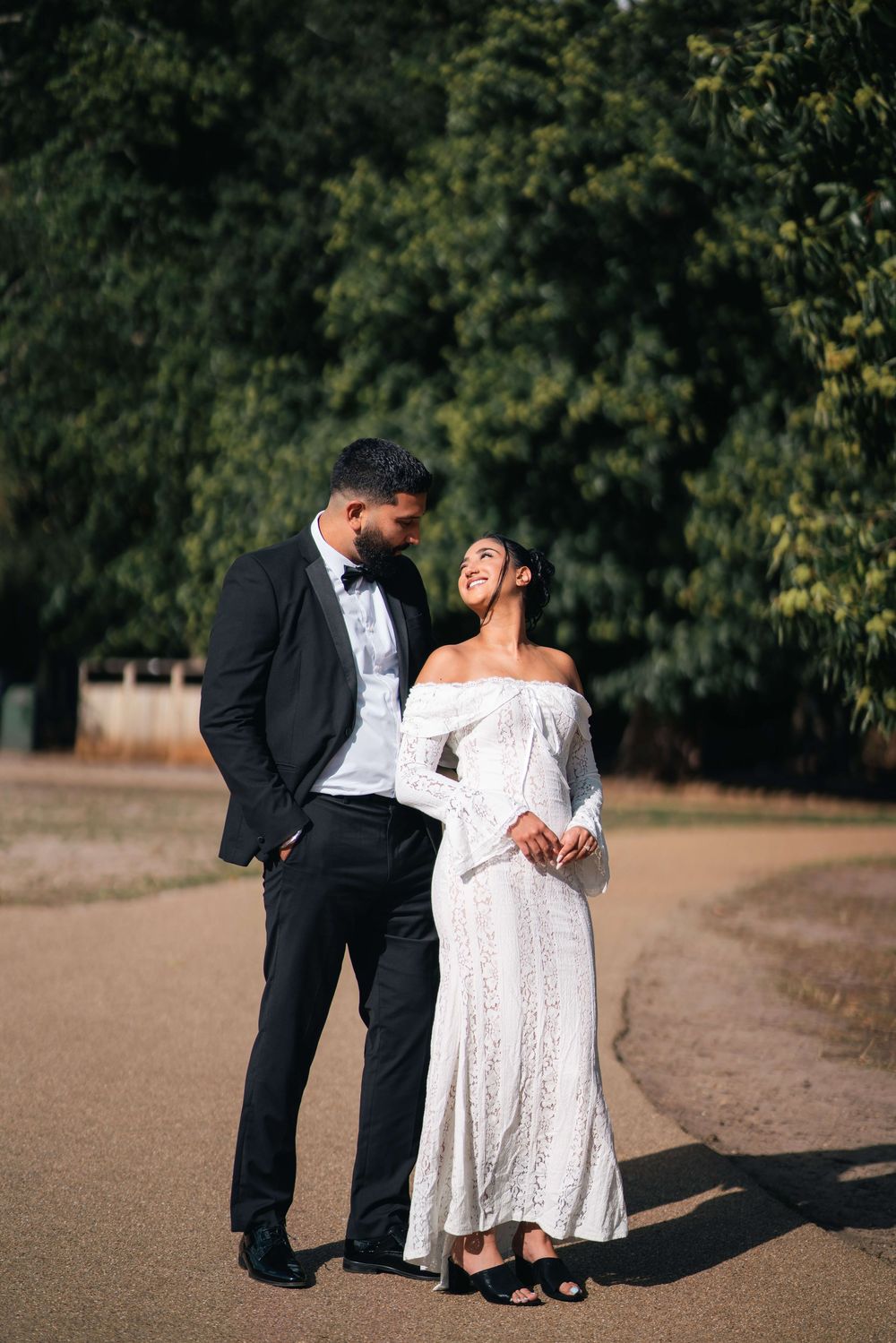 Smiling couple in formal wedding attire, a groom in a black tuxedo and a bride in an off-shoulder lace gown, pose on a park path.