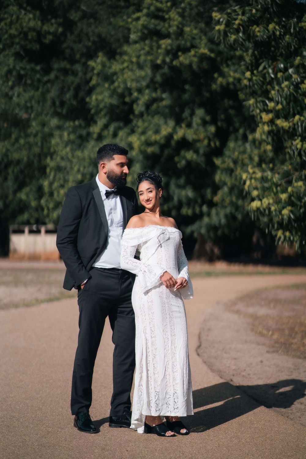 Couple in formal wear, a man in a black tuxedo with bow tie and a woman in a white off-shoulder lace gown, posing on a path near trees.