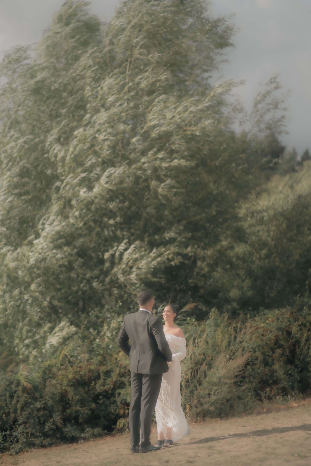 Bride in a white off-the-shoulder gown faces a groom in a gray suit beneath wind-swept willow trees during an outdoor wedding portrait.