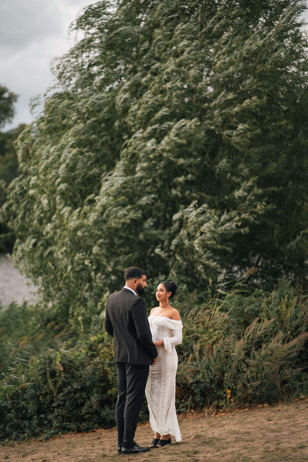 Couple in formal wedding attire stand facing each other on a grassy path beside windswept green trees under a cloudy sky.