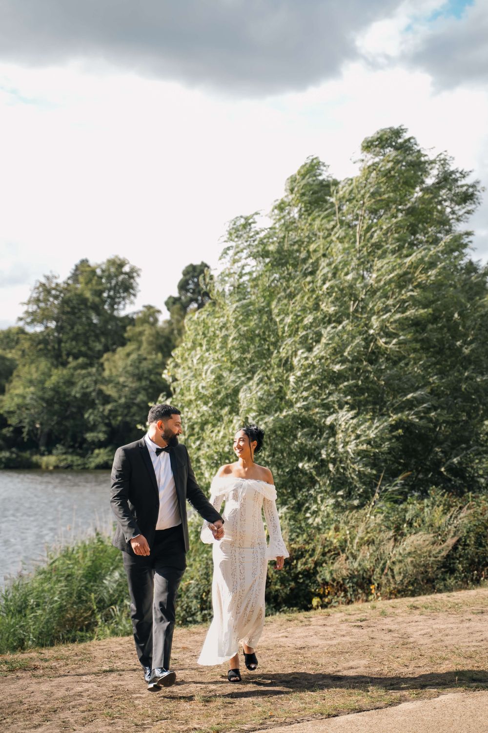 Bride in a white off-shoulder dress and groom in a black suit and bow tie walk hand in hand by a lake on a windy day.