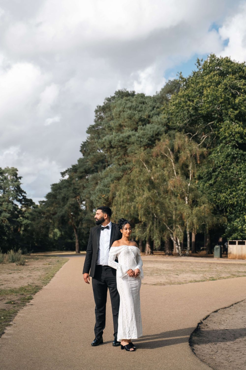 Couple in formal wedding attire, a man in a black tuxedo and a woman in a white off-shoulder gown, pose on a park path beneath tall trees.