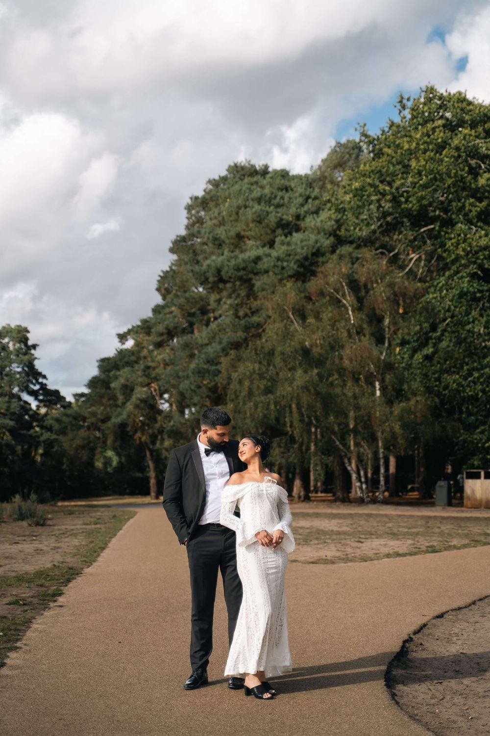 Couple in formal wedding attire, a groom in a tuxedo and bride in a white off-shoulder gown, embracing on a path near tall trees.