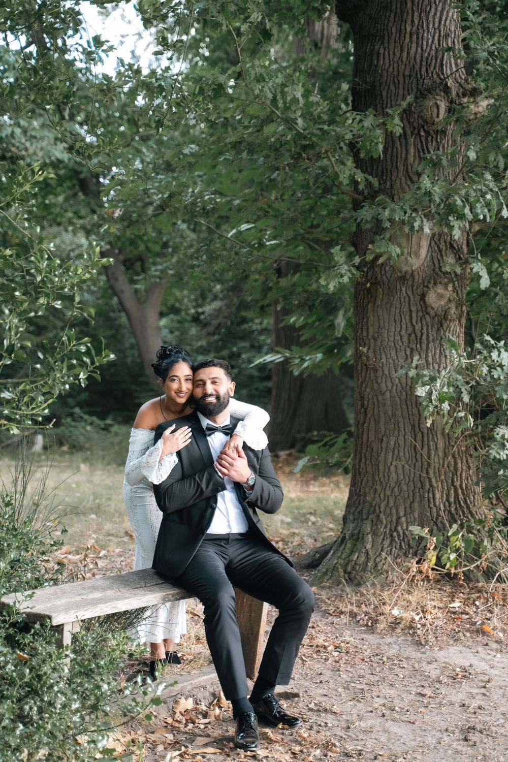 Couple in formal wedding attire embracing on a wooden bench beneath a large tree in a sunlit park, the bride hugging the groom from behind.