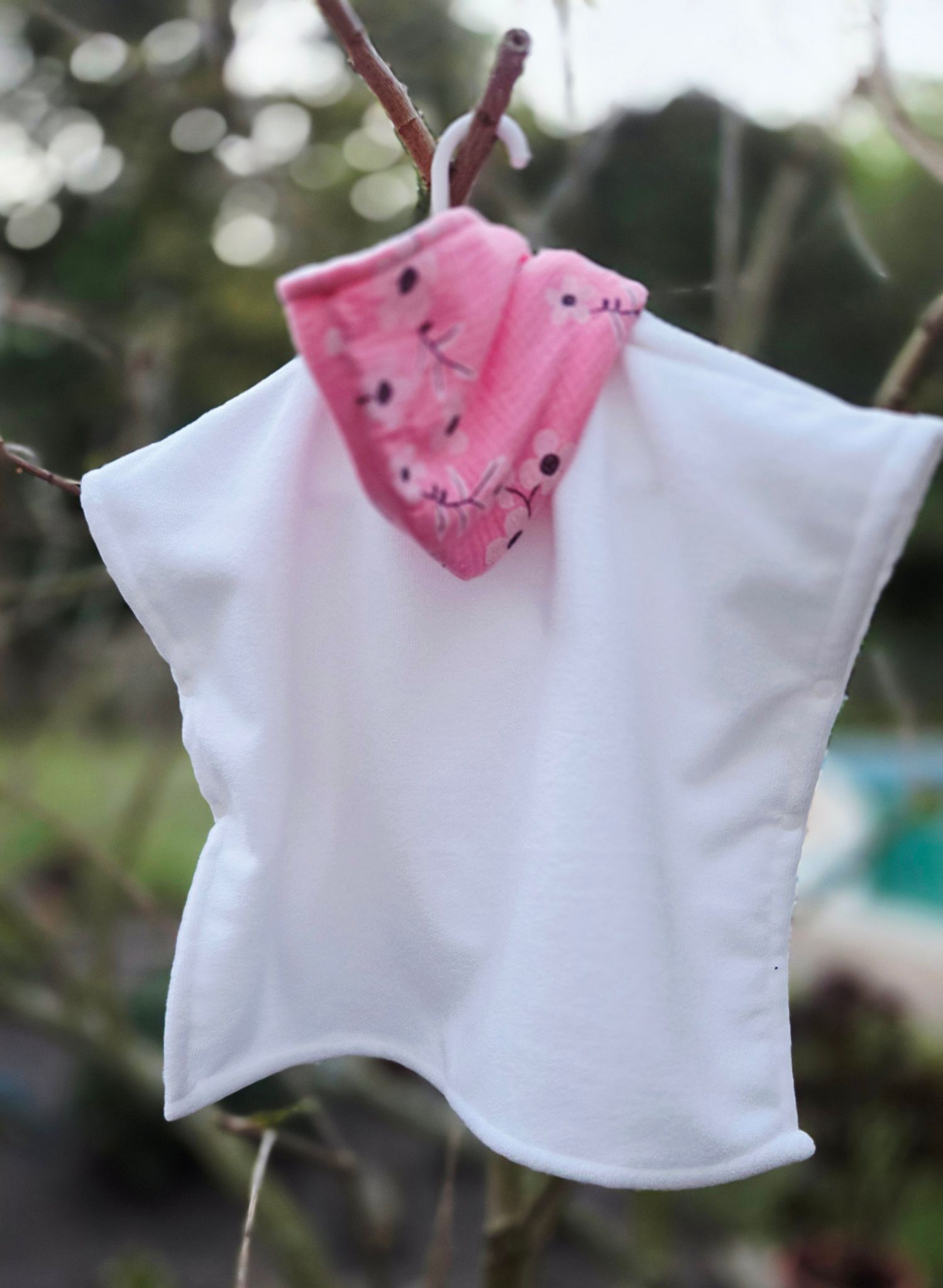 White baby poncho with pink floral collar hanging on a branch outdoors.
