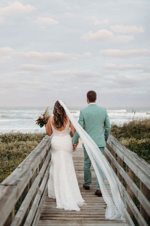 Bride and groom walking hand-in-hand down a wooden boardwalk toward the ocean.