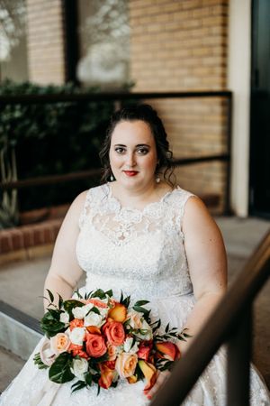 Plus-size bride in lace wedding gown holds vibrant orange and white rose bouquet on steps.