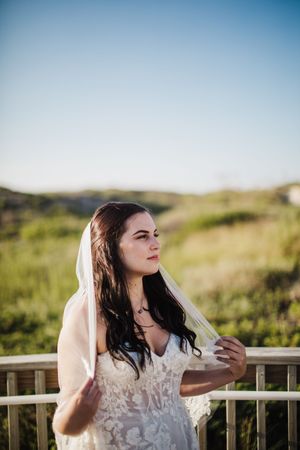 Dark-haired bride in lace gown and veil standing on a boardwalk in a sunny field.