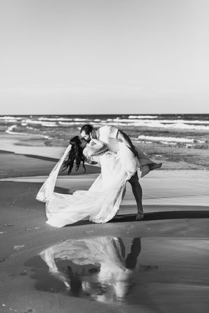 Black and white photo of groom dipping bride on beach shoreline with stunning reflection in wet sand.