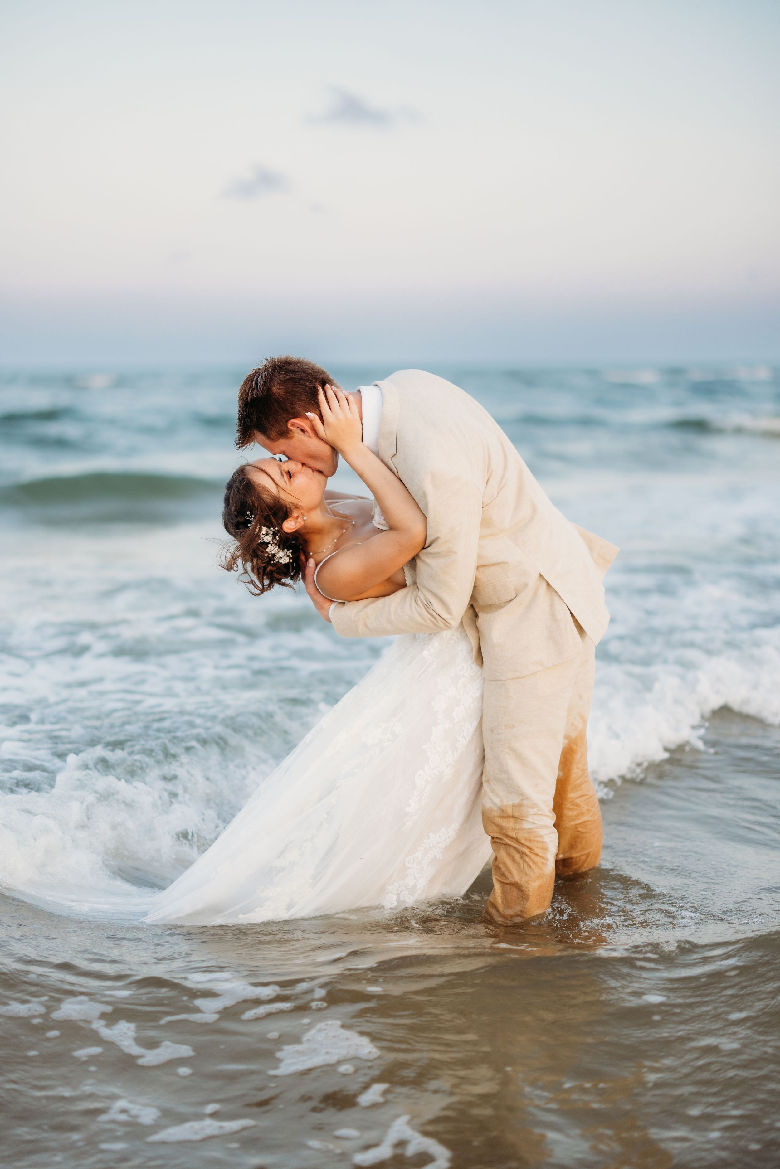 Groom dipping bride for a kiss while standing in ocean waves at a beach wedding.