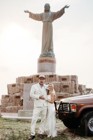 Couple in white wedding attire posing before a Christ statue beside a vintage SUV.