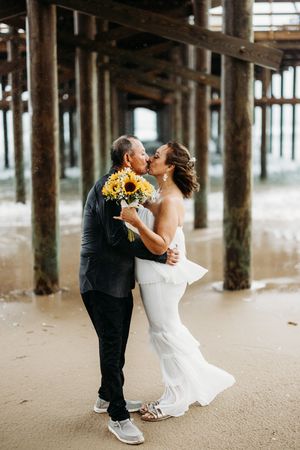 Couple kissing under a pier with bride holding a vibrant sunflower bouquet at beach wedding.