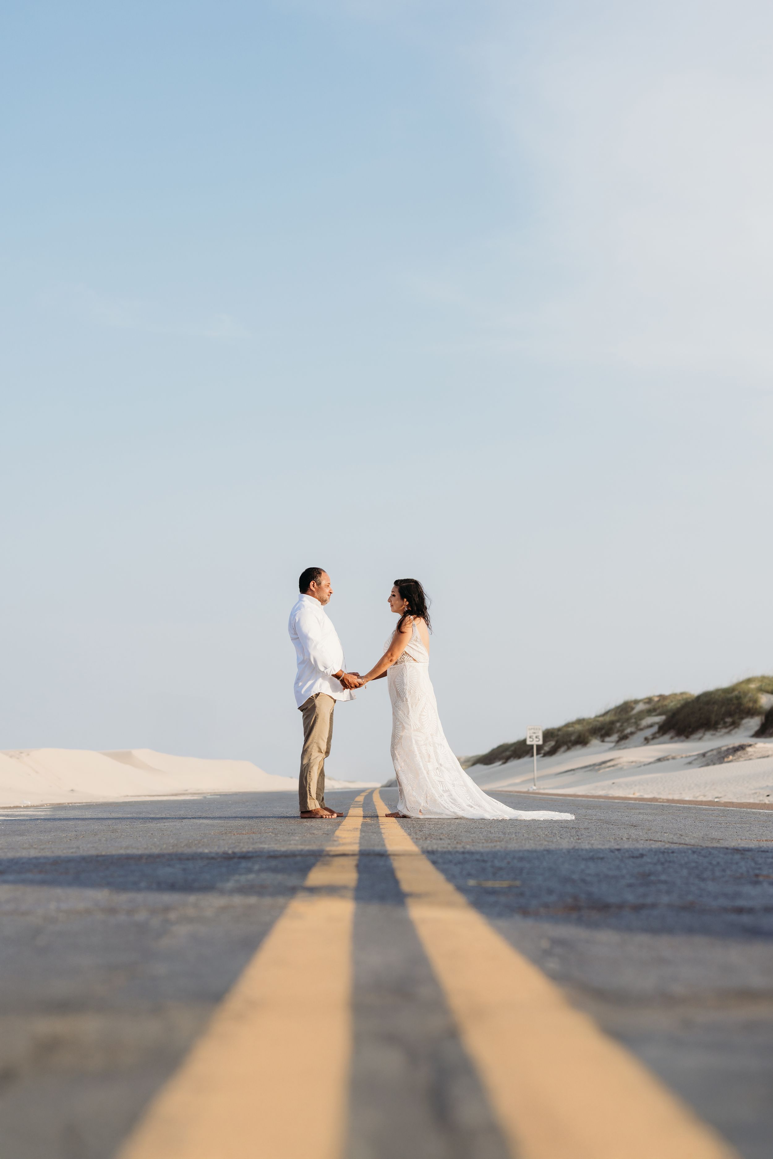 Bride and groom hold hands on empty road surrounded by sand dunes under clear blue sky.