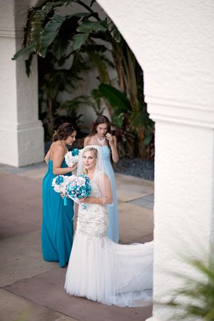 Bride in beaded gown holds teal bouquet with bridesmaids in turquoise dresses under arched walkway.