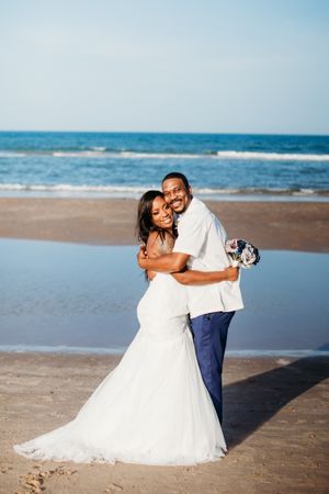 Happy newlywed couple embraces on sunny beach, bride in mermaid gown, groom in white shirt and navy pants.