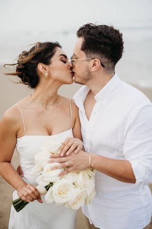 Bride and groom sharing a kiss on the beach while holding a white rose bouquet.
