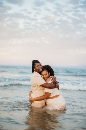 Two Black women in beige outfits embracing on a beach at sunset in the ocean waves.