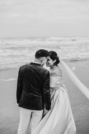 Black and white photo of bride and groom sharing a tender moment on a beach shoreline.