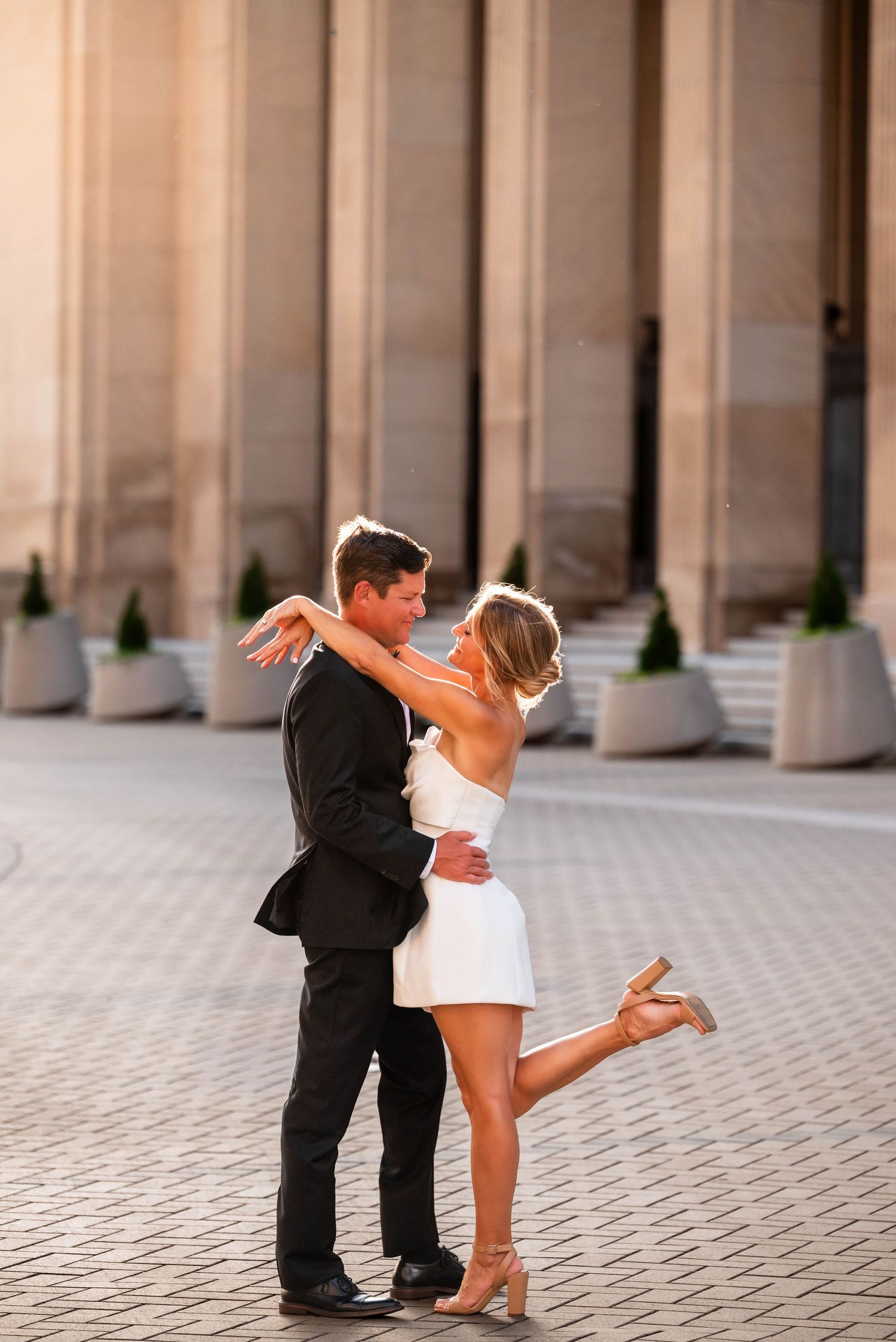 Joyful couple in wedding attire embracing on a city street, groom lifting bride in white dress as both laugh.