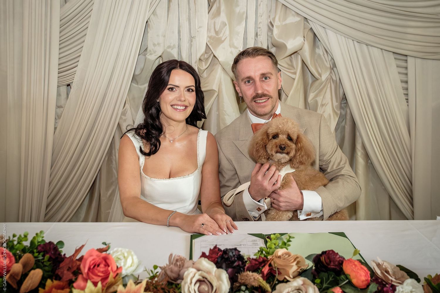 Happy couple in wedding attire smiling with a fluffy dog at a floral-decorated reception table.