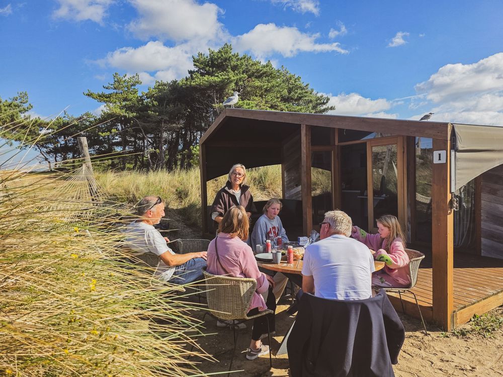 Family gathering at outdoor picnic table beside a wooden beach cabin surrounded by dunes and pine trees on a sunny day.