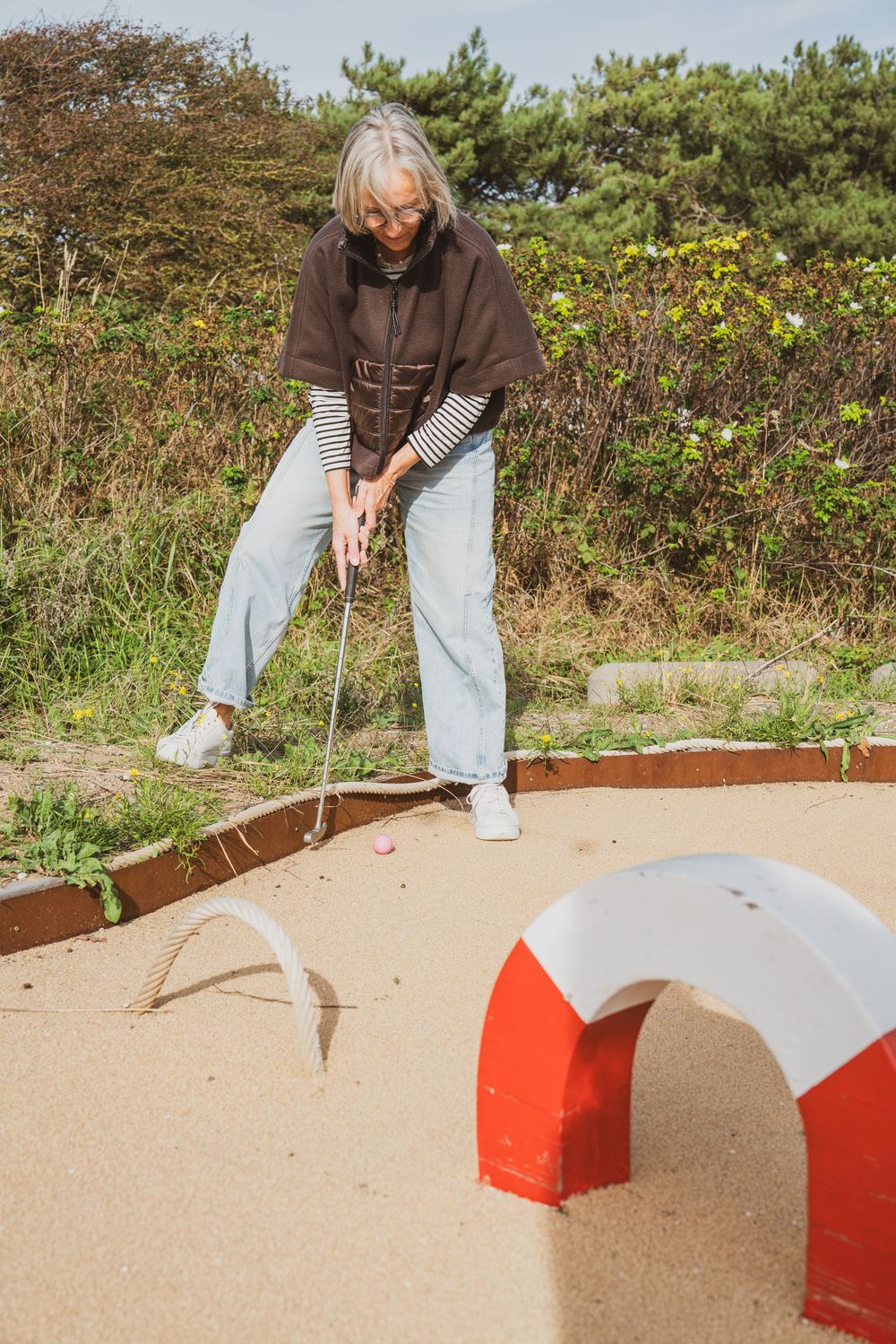 Senior woman with gray hair playing mini golf outdoors, putting near a red and white arch obstacle on a sandy course.